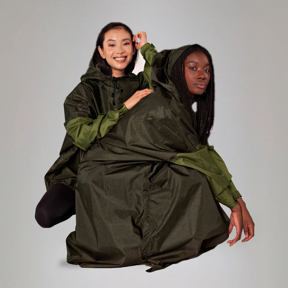Two people wearing a large green and black poncho against a plain background